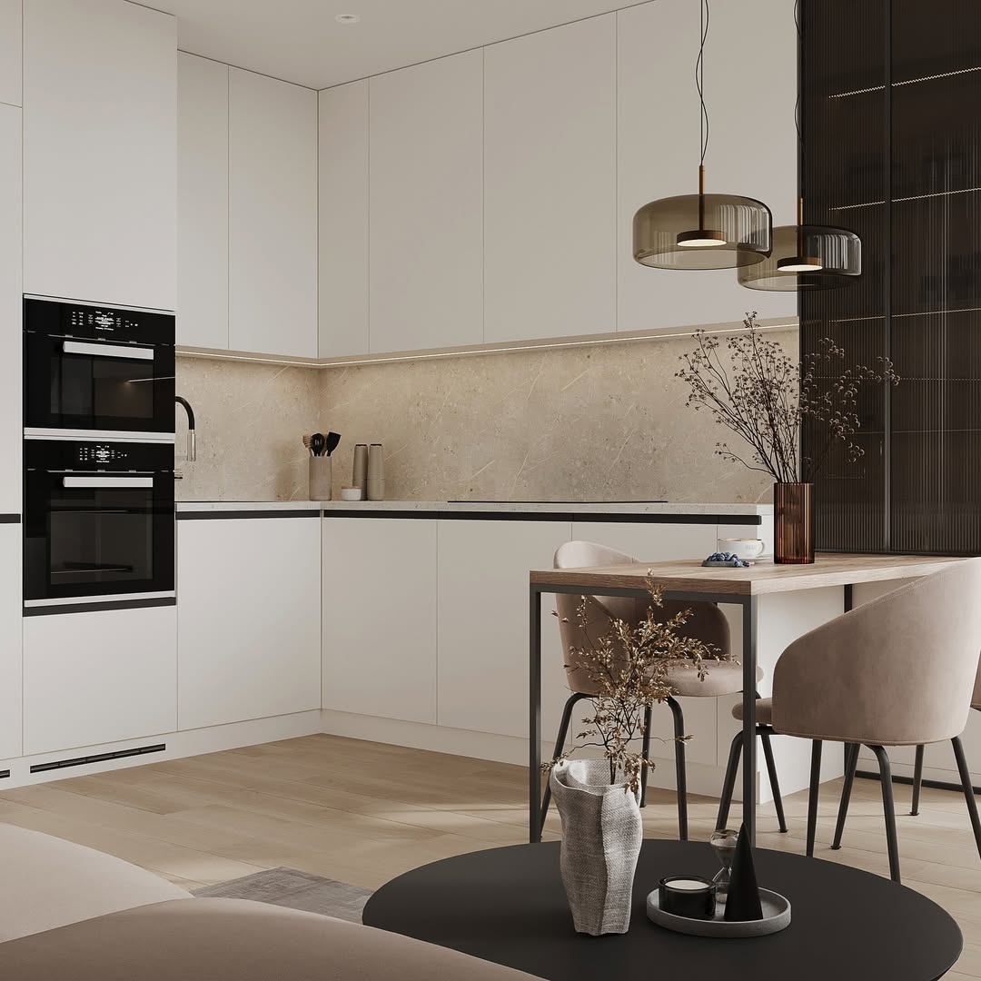 Modern kitchen with sleek white cabinets, beige stone backsplash, built-in black ovens, and a small dining table with blush velvet chairs and pendant lights above.